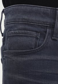 Mörka jeans med en närbild av midjan och fickan. Texturerat tyg med synliga sömmar och en klassisk passform.