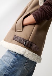 Gilet beige con rifiniture in shearling e maniche a coste marroni, dotato di una tasca laterale e di una cintura marrone scura in vita. Materiale testurizzato.