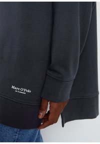 Sudadera gris oscura con puños de canalé y aberturas laterales, que presenta un detalle de logo blanco en el dobladillo. Textura de tela suave.