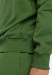 Sudadera verde con puños y dobladillo acanalados, que presenta una textura suave y un diseño sin costuras en los hombros, mostrada parcialmente en un antebrazo.