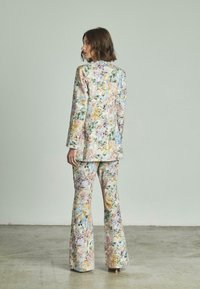 Completo floreale con giacca aderente e pantaloni a zampa in colori pastello, realizzato in un tessuto testurizzato che presenta motivi viola, giallo e verde.