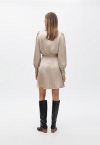 Vestido de satén beige con mangas largas y abullonadas, y cintura ceñida. Combinado con botas negras hasta la rodilla y sutiles detalles de botones dorados.