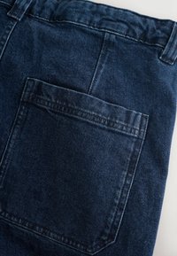 Jeans de mezclilla azul oscuro con un solo bolsillo trasero, costuras visibles y un diseño clásico de cinturilla. La tela tiene una superficie ligeramente texturada.
