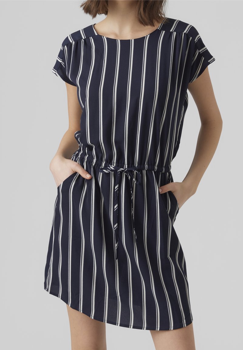Femme portant une robe bleu marine à rayures verticales blanches, manches courtes, taille cintrée avec lien, mains dans les poches.