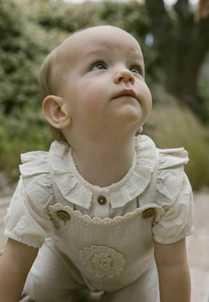 Salopette en lin de couleur claire avec des détails de broderie florale, col à volants et boutons en bois, portée sur une chemise à manches courtes blanche.