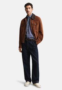 Mężczyzna w brązowej zamszowej kurtce, niebieskiej koszuli na guziki, ciemnych dżinsach, plecionym pasku i białych trampkach, stoi z jedną ręką w kieszeni.