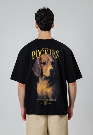 Mand iført en sort T-shirt med et stort realistisk hundeportæt og gul tekst, der siger "House of Pockies" på ryggen.