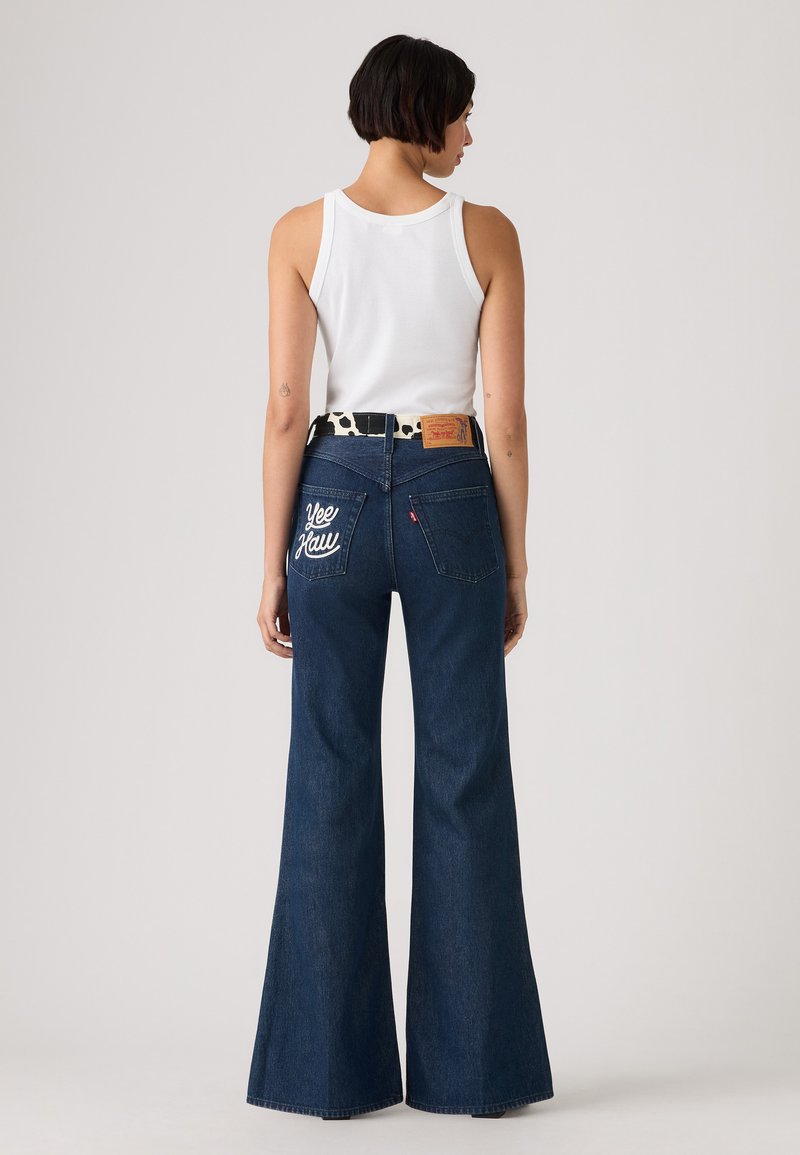 Mørkeblå udbrede denim jeans med hvid broderet tekst "Yee Haw" på baglommerne, udstyret med et mønstret sort-hvidt bælte.