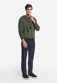 Grön texturerad tröja med en scarf över axlarna, kombinerad med mörka jeans och svarta skor med vita sulor.