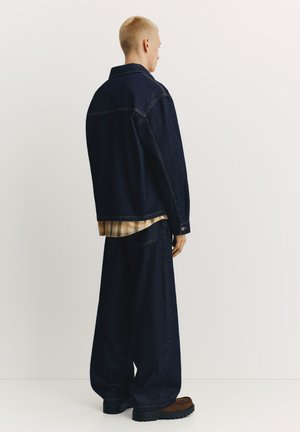 Veste en jean et jean à jambes larges bleu foncé, superposés sur une chemise à carreaux jaune. Présente des coutures visibles et un tissu texturé. Chaussures noires épaisses.