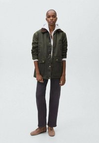 Vihreä kanvastakki ruskealla kauluksella, kaksi etutaskua, harmaan hupparin päällä, yhdistetty tummiin housuihin ja vaaleanruskeisiin kenkiin.