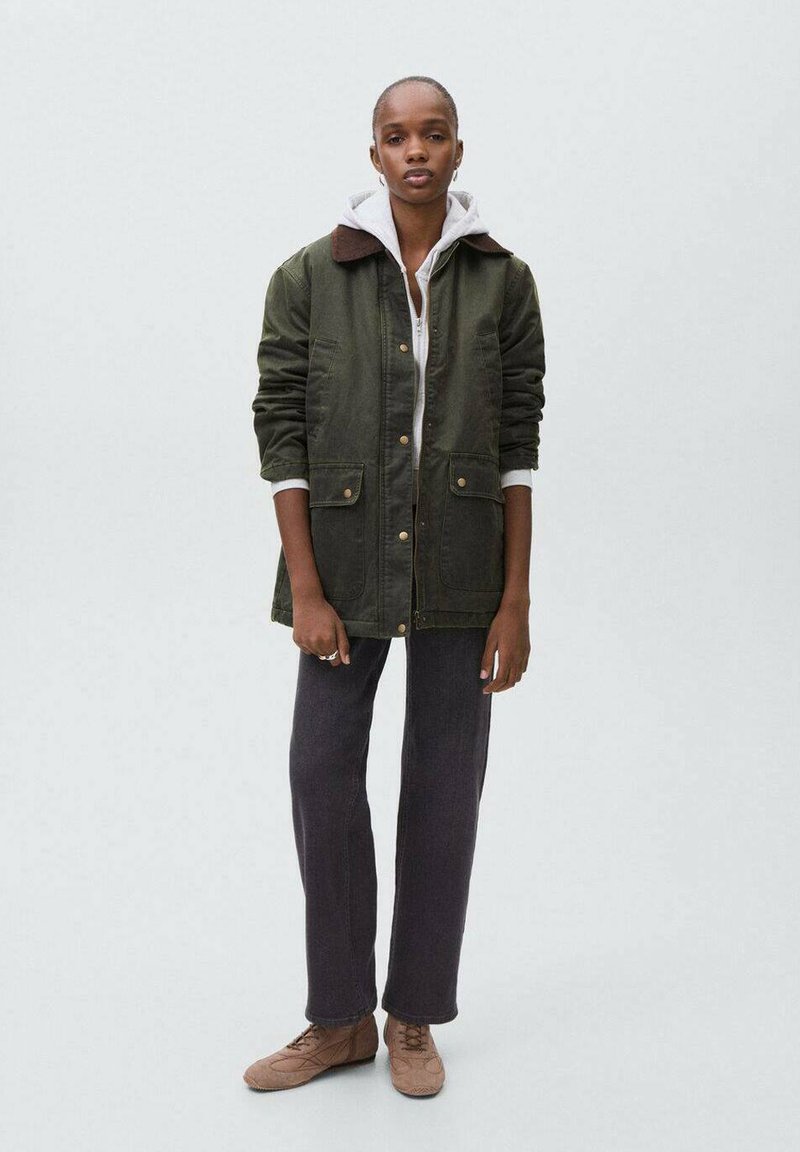 Vihreä kanvastakki ruskealla kauluksella, kaksi etutaskua, harmaan hupparin päällä, yhdistetty tummiin housuihin ja vaaleanruskeisiin kenkiin.