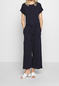 Marineblauer Overall mit kurzen Ärmeln, einer Fronttasche, weiten Beinen und einem gebundenen Gürtel in der Taille; kombiniert mit weißen, offenen Mules.