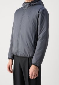 Mann in einer grauen Zip-Jacke mit Kapuze, elastischen Bündchen und Taschen, kombiniert mit schwarzen Hosen vor einem schlichten Hintergrund.
