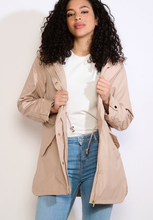 Donna con capelli ricci che indossa una giacca beige con cappuccio sopra una maglietta bianca e jeans blu, tenendo aperta la giacca.