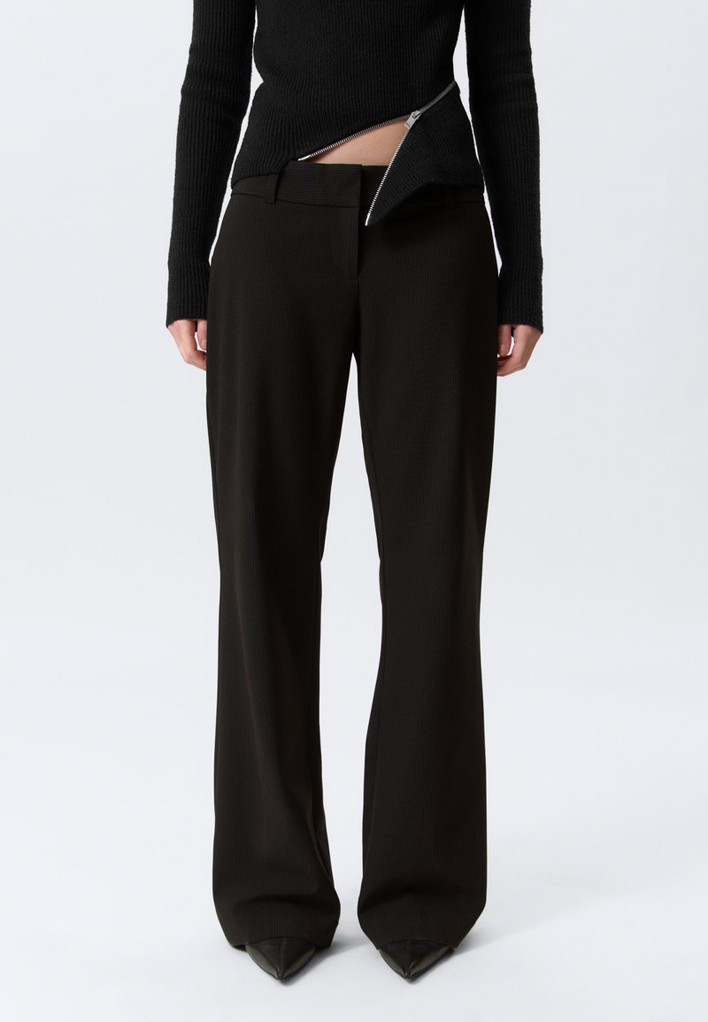 Personne portant un pantalon noir à jambes larges et un pull côtelé noir avec une fermeture éclair asymétrique dévoilant le bas de la taille.