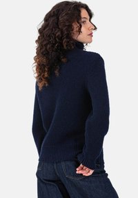 Pull à col roulé bleu marine en maille douce et texturée, avec poignets et ourlet côtelés, porté avec un jean bleu foncé sur un fond uni.