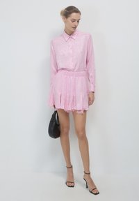 Abito camicia rosa in tessuto texture, con bottoni sul davanti e vita elastica, caratterizzato da una gonna svasata e una borsa nera. Tacchi neri con cinturini.