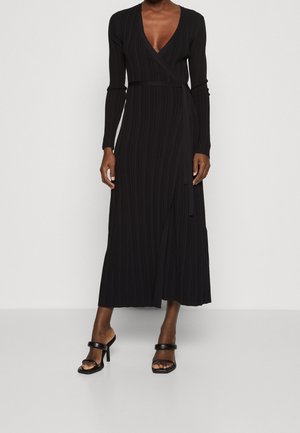 Kobieta ubrana w czarną, plisowaną sukienkę kopertową z długim rękawem i dekoltem w serek oraz czarne sandały na wysokim obcasie, stojąca na jednolitym tle.