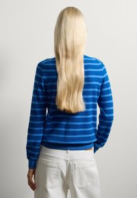 Maglione a righe blu con colletto rotondo e polsini a coste, abbinato a pantaloni bianchi. Texture liscia, silhouette aderente e design casual.