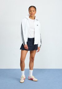 Giacca bianca con cerniera e strisce nere, t-shirt bianca, gonna blu navy e sneakers pastello con dettagli rossi. Capelli intrecciati e orecchini a cerchio.