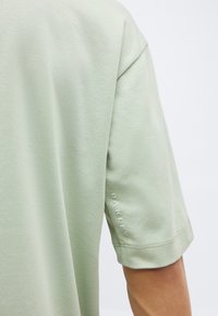 Πράσινο ανοιχτό κοντομάνικο πουκάμισο με το λογότυπο "Oakley" ανάγλυφο στο μανίκι, φορεμένο από ένα άτομο μπροστά σε απλό λευκό φόντο.