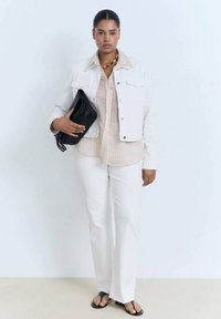White denim jacket over a light striped shirt, paired with white pants and black clutch. Model wears black flip-flops and wooden bead necklace.