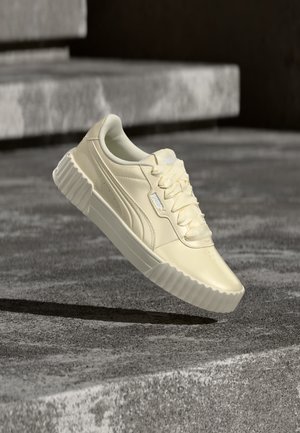 Off-white Puma sneaker med tyk ribbet sål og brede satin snørebånd vist over en betongoverflade og trin.