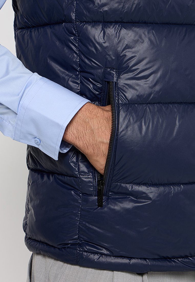 Gilet rembourré bleu marine avec un design matelassé, doté d'une poche zippée sur le côté gauche et d'une texture lisse et brillante. Chemise à manches longues bleu clair en dessous.