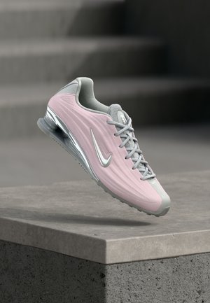 Lichtroze en grijze sportschoen met zilveren Nike-swoosh, zwevend boven een betonnen richel met trappen op de achtergrond.