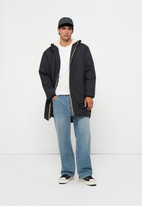 Veste à capuche noire et isolée avec doublure beige, accents de zip jaune, portée sur un T-shirt blanc, associée à un jean bleu clair et des baskets.