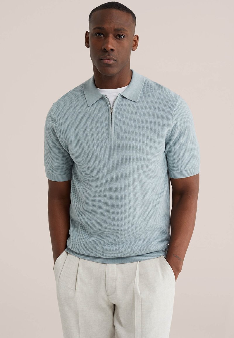 Muž v světle modrém pleteném polo tričku s krátkým rukávem a zipem, v kombinaci se světle béžovými plisovanými kalhotami, stojící s rukama v kapsách.
