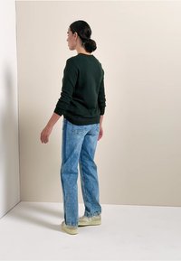 Tmavě zelený svetr, volný střih, spárovaný s světle modrými kalhotami širokého střihu. Na sobě má světlé tenisky, stojící proti neutrálnímu pozadí.