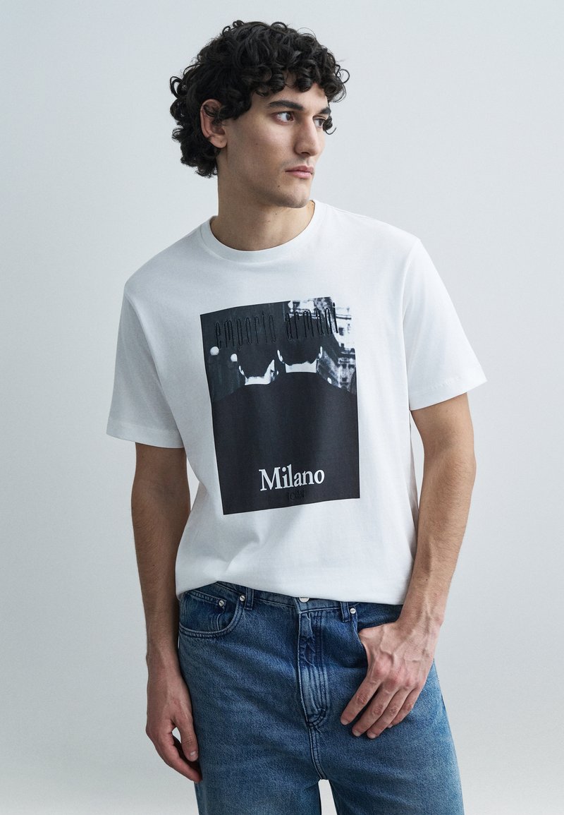 Biele bavlnené tričko s čiernou grafickou potlačou s textom "Emporio Armani" a "Milano" v dolnej časti. Nosené s modrými rifľami.