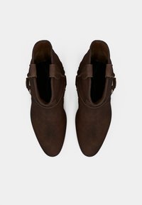 Ζευγάρι μπεζ σκούρων δερμάτινων μποτών αστραγάλου με λουριά και αγκράφες, φωτογραφημένα από πάνω σε απλό φωτεινό φόντο.