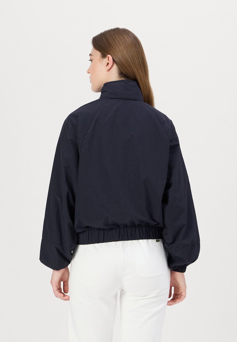 Veste courte bleu marine avec un col montant, une taille élastique et des manches longues. Présente une texture lisse et des détails de design subtils.