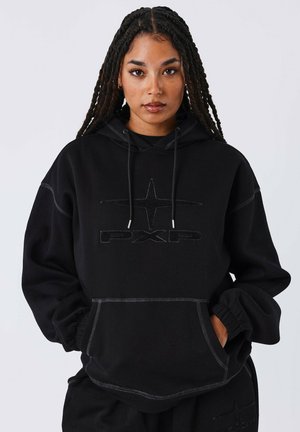 Vrouw met gevlochten haar, gekleed in een zwarte hoodie met een sterrenlogo en de tekst "PXP", met haar handen in de voorzak, tegen een eenvoudige achtergrond.