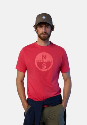 Uomo con barba che indossa un berretto oliva, una maglietta rossa con logo North Sails, pantaloni verdi e una maglia blu navy legata in vita su sfondo uniforme.