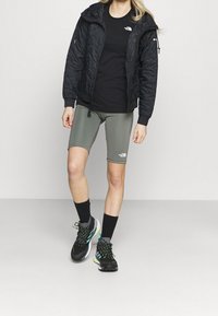 Czarna pikowana kurtka, czarna koszulka, zielone szorty, czarne i szare buty sportowe z neonowymi akcentami, noszone z czarnymi skarpetami.