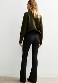 Vrouw met blond haar in een olijfgroen trui en zwarte aansluitende flare broeken die met haar rug naar de kijker tegen een witte muur staat.