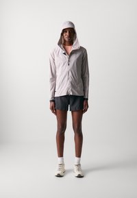 Giacca leggera grigia chiaro con cappuccio, zip frontale e dettagli scuri, abbinata a pantaloni corti neri e scarpe da ginnastica color crema.