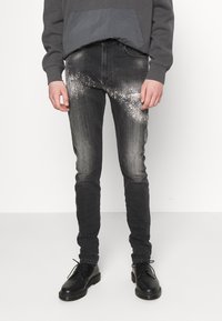 Vaqueros de mezclilla negra con un ajuste slim, que presentan un patrón de salpicaduras grises texturizadas en la parte delantera. Combinados con una sudadera gris oscuro y zapatos negros.