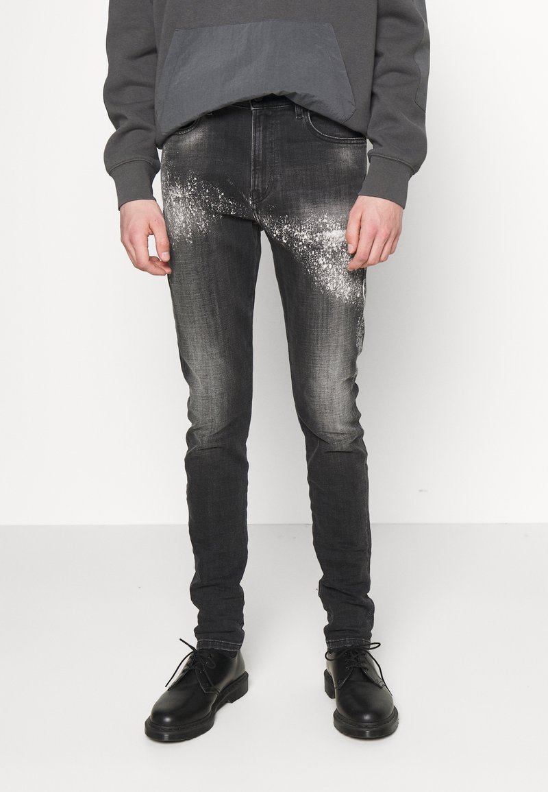 Vaqueros de mezclilla negra con un ajuste slim, que presentan un patrón de salpicaduras grises texturizadas en la parte delantera. Combinados con una sudadera gris oscuro y zapatos negros.