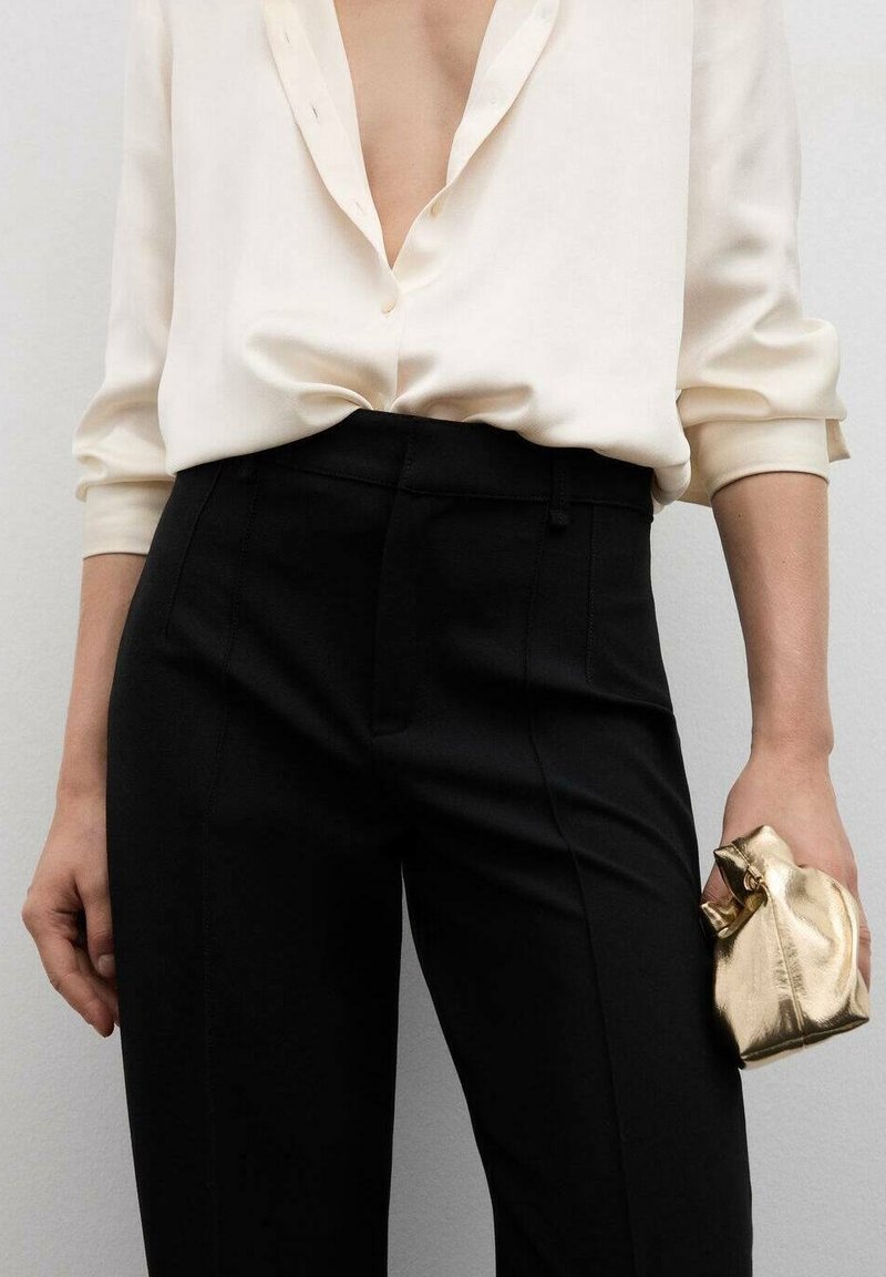 Blouse en soie blanche avec un profond décolleté en V et des manches retroussées, associée à un pantalon noir ajusté taille haute. La main tient un petit sac métallique doré.