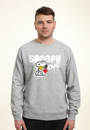 Młody mężczyzna w szarej bluzie z nadrukiem Snoopy'ego obejmującego Woodstocka i czerwonym sercem na przodzie, stoi na jasnym jednolitym tle.