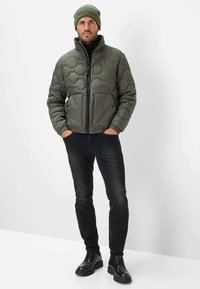 Polstret olivengrøn jakke med høj krave, lynlås lukning og to forlommer, parret med sorte jeans og sorte sko. Beanie-hat inkluderet.