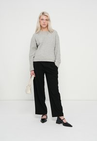 Szürke kötött pulóver fekete, bőszárú nadrággal párosítva. A modell egy kis krémszínű táskát tart. Fekete, hegyes orrú cipő teszi teljessé az outfitet.