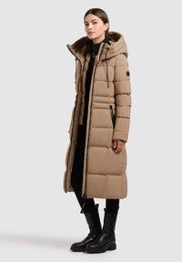 Beige színű párnázott hosszú kabát kapucnival, elöl cipzárral, fekete részletekkel és texturált felülettel. Fekete ruházat és csizma fölött viselve.