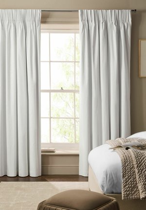 Weiße plissierte Vorhänge, die ein großes Fenster mit Sonnenlicht und Bäumen draußen einrahmen, neben einem Bett mit einer beigen Steppdecke und einer geöffneten Zeitschrift.