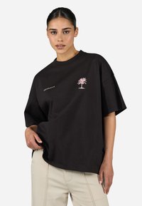 Camiseta de algodón negra de corte oversize con mangas cortas, que presenta un pequeño bordado de una palmera rosa y el texto "PEGADOR" en el frente.